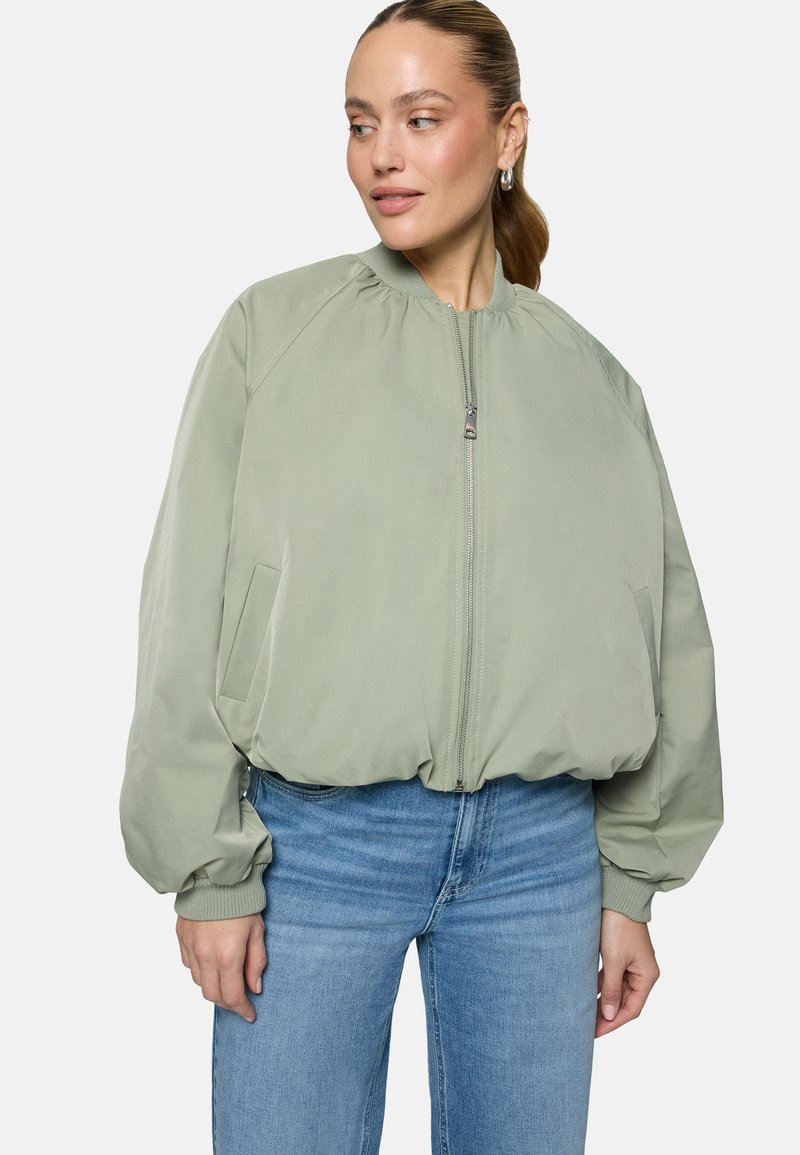 Femme portant une veste bomber zippée vert clair et un jean bleu, regardant sur le côté, les cheveux attachés, devant un fond uni.