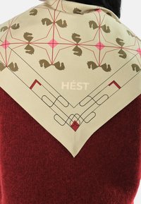 Silke skjerf i lys beige med et geometrisk mønster av brune hester og rosa detaljer, med dekorative svarte og røde kanter. Inkludert teksten "HÉST".