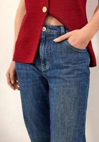 Gilet rosso testurizzato con un bottone dorato, abbinato a jeans in denim blu lavati con tasca laterale e cuciture a contrasto.