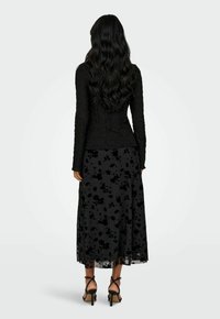 Donna con lunghi capelli neri mossi, che indossa un top a maniche lunghe nero con texture, una gonna midi nera a fiori e tacchi alti neri con cinghie, in piedi.