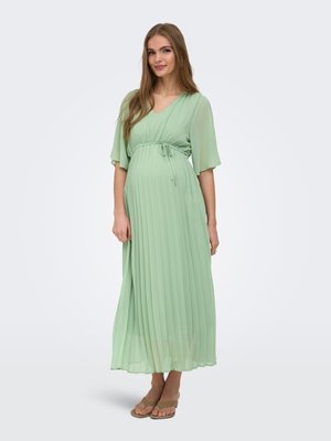 Femeie însărcinată cu păr lung, îmbrăcată într-o rochie maxi plisată de culoare verde deschis și sandale bej, stând în fața unui fundal simplu.