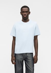 Helehall sinine puuvillane T-särk ümar kaeluse ja lühikeste varrukatega. Rinnal on valgete tähtedega peen tekst. Sobitatud tumedate teksapükstega.