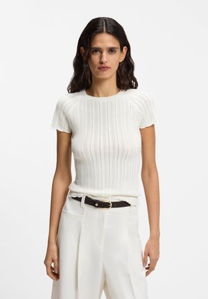 Femme aux longs cheveux foncés portant un haut en maille blanc à manches courtes et un pantalon blanc taille haute avec une ceinture noire.