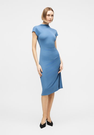 Γυναίκα με στενή εφαρμοστή μπλε μίντι φόρεμα με κοντά μανίκια και μαύρες γόβες με μυτερή μύτη, στέκεται μπροστά σε απλό φωτεινό φόντο.