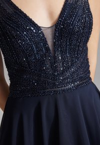 Laivansinis mekko, jossa on syvä V-kaula-aukko, jossa on monimutkaisia helmi koristeluita ja laskostuva hame, valmistettu sileästä kankaasta.