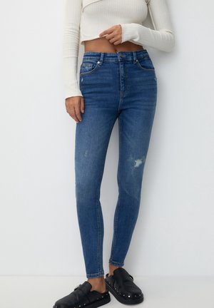 Femme portant un jean skinny bleu taille haute avec légère usure, un pull court blanc côtelé et des chaussures noires à enfiler avec des clous.