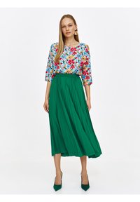 Bluzka w kwiatowy wzór na niebieskim tle, zestawiona z plisowaną zieloną spódnicą maxi; modelka w zielonych szpilkach stoi na białym tle.