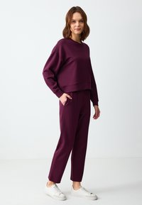 Vrouw die staat in een bijpassende donkerpaarse sweatshirt en broek met witte sneakers tegen een effen witte achtergrond.