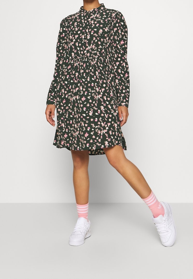 Langärmliges, knöpfbares Kleid in Dunkelgrün mit rosa und weißen Blumenmuster. Kombiniert mit weißen Sneakers und rosa gestreiften Socken.
