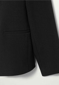 Manche de veste de costume noire et poche avant inférieure avec une couture nette sur fond clair uni.