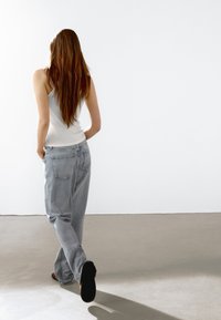 Lichtgrijze straight-leg jeans met achterzakken en een witte tanktop. De figuur loopt weg tegen een effen witte achtergrond.