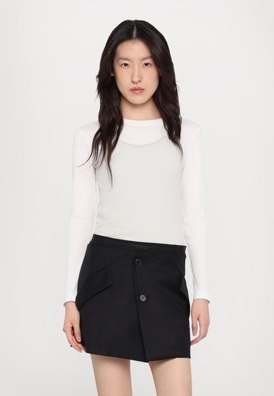 Balts garais piedurkņu tops ar rievojuma tekstūru, kas valkāts zem melnas apvītu svārku, kas izceļas ar priekšējo pogu un unikālu kabatu detaļu.