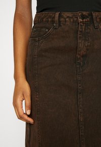 Bras et main gauches reposant à côté d'une jupe en jean marron taille haute avec poche avant visible et passants de ceinture.