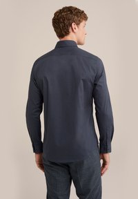 Chemise bleu marine à manches longues avec un col rigide, texture lisse et poignets avec boutons, vue de dos sur un fond neutre.