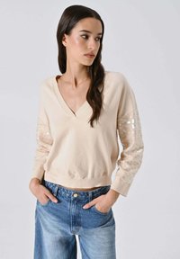Maglione beige a scollo a V con maniche testurizzate impreziosite da paillettes. Polsini e orlo a coste; abbinato a jeans di denim blu a vita alta.
