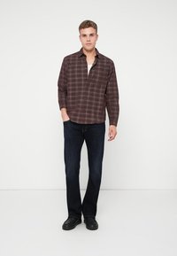 Brun rutete skjorte med knapper og et avslappet snitt, sammen med mørke denimjeans. Modellen står foran en nøytral bakgrunn med svarte sko.