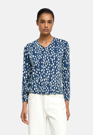 Kobieta ubrana w niebieski sweter z dekoltem w serek z białymi owalnymi plamami i białe spodnie, stojąca na tle jednolitego jasnego tła.