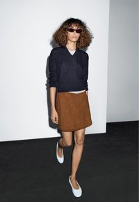 Schwarzer Pullover mit Struktur, brauner Wildlederrock und weiße Slip-On-Schuhe. Das Model trägt braun getönte Sonnenbrillen und steht vor einem neutralen Hintergrund.