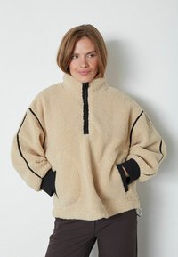 Bež flis pulover s polu-zip prednjim dijelom, crnim detaljima, oversized krojem i bočnim džepovima. Teksturalna i meka tkanina, opuštene rukave.