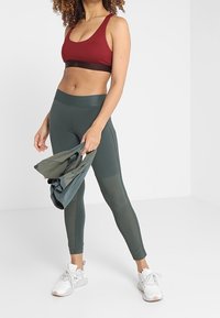 Bra sportivo rosso con una fascia scura, abbinato a leggings verdi a vita alta. La modella tiene un top coordinato, realizzato in un materiale morbido e elastico.