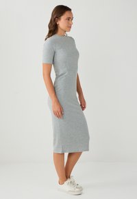 Vrouw die zijwaarts staat in een aansluitende, geribde, grijze midi-jurk met korte mouwen en witte sportschoenen tegen een effen achtergrond.