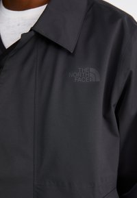 Manteau léger noir avec un col pointe, présentant un imprimé logo mat "The North Face" sur la poitrine, texture lisse et détails de coutures.