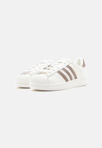 Λευκά αθλητικά παπούτσια με υφή στο μπροστινό μέρος, που διαθέτουν τρεις καφέ και γκρι ρίγες, επίπεδα κορδόνια και λείο δερμάτινο επάνω μέρος.