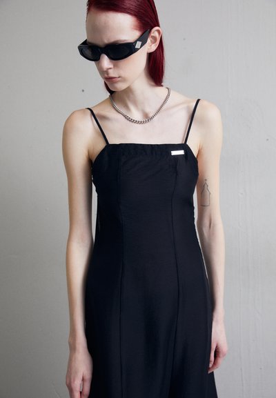 Čierne šaty na ramenách so strmým strihom, s malým strieborným akcentom na hrudi. Model nosí čierne slnečné okuliare a strieborný náhrdelník.