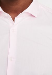 Camicia button-up di un delicato rosa chiaro, con una texture liscia, colletto classico e una leggera lucentezza. Presenta bottoni in plastica trasparente lungo la pattina.