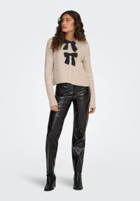 Beige neulejakku, jossa kaksi mustaa rusettiyksityiskohtaa, yhdistettynä mustiin kiiltäviin nahkalegginsseihin ja mustiin nilkkureihin.