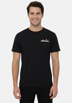 Homme souriant portant un t-shirt noir avec le logo ellesse, debout les bras détendus le long du corps devant un fond clair uni.