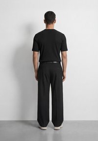 Άνδρας με μαύρο κοντομάνικο πουκάμισο, μαύρο παντελόνι με ζώνη και λευκά αθλητικά παπούτσια, με την πλάτη προς τον θεατή, μπροστά σε λευκό τοίχο.