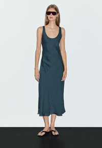 Abito midi senza maniche, elegante, di un verde scuro, realizzato in un tessuto morbido e fluido con una sottile texture; indossato con sandali neri piatti e occhiali da sole oversize.