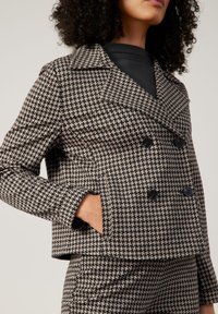 Doppelreihige Jacke in Schwarz-Beige mit Hahnentritt-Muster, spitzem Kragen, Vordertaschen und schwarzen Knöpfen, strukturiertem Stoff.