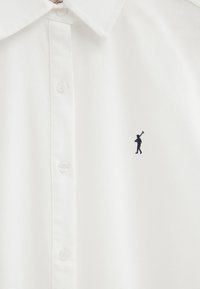 Chemise blanche à boutons avec col pointu et petite figure noire brodée tenant une hache sur le côté gauche de la poitrine.