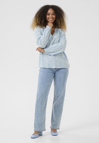 Helle blaue Bluse mit floralem Muster und elastischen Bündchen, getragen mit hellblauen geraden Jeans und passenden hohen Schuhen, vor einem weißen Hintergrund.
