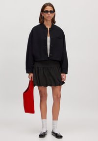 Zwarte cropped jasje met ronde kraag en knoopsluiting, gecombineerd met een zwarte rokken met ruches en een rode tote bag. Model draagt een zonnebril.