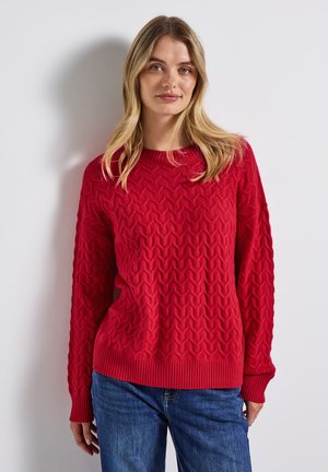 Rode gebreide trui met een gestructureerd zigzagpatroon, ribbelkraag, en lange mouwen, gecombineerd met blauwe jeans.