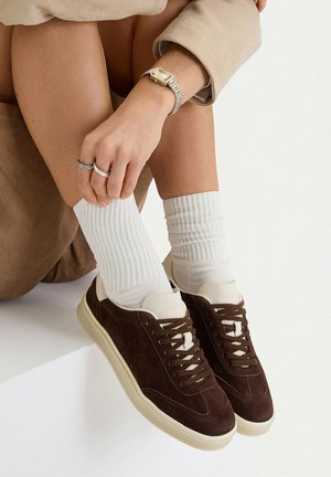 Piernas cruzadas con calcetines blancos acanalados y zapatillas de ante marrón, mano con anillos de plata y reloj dorado apoyada en la rodilla, manga de abrigo beige visible.