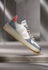 White and gray sneaker with beige logo, gum sole, and red heel accent, suspended above a stone surface with a blurred background.
