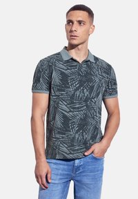 Camisa polo de manga corta en verde oscuro con un patrón de hojas, que presenta una tela suave, un cuello y un ajuste holgado. Usada con unos jeans azul claro.