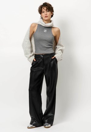 Grijze mouwloze top met hoge kraag en logo, gecombineerd met zwarte wijde nep leren broek. Model draagt zilveren sneakers en houdt haar handen in de zakken.