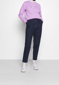 Lila sweatshirt med "TOMMY JEANS" logotyp, ihop med högmidjade mörkblå jeansbyxor och ljusgrå sportiga sneakers.