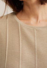 Beige stickad tröja med rund halsringning och vertikala sömdetaljer. Mjuk textur med en lätt lyster och ribbat detaljer vid kragen.