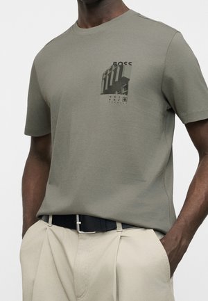 Man draagt een olijfgroen BOSS t-shirt met een zwarte afbeelding en beige broek met een zwarte riem, handen in de zakken.