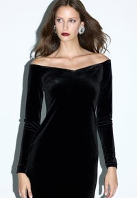 Vestido de terciopelo negro sin hombros con mangas largas y silueta ajustada, que presenta un escote en V y una textura suave.