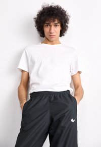 Λευκό μπλουζάκι από βαμβάκι με στρογγυλή λαιμόκοψη, συνδυασμένο με μαύρα παντελόνια φόρμας με ελαστική μέση και ένα μικρό λογότυπο Adidas στον αριστερό μηρό.