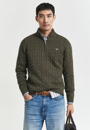 Mann iført olivengrønn kabelstrikket genser med halv glidelås, blå jeans, brunt belte, holder mørk brun skinnveske mot ensfarget bakgrunn.