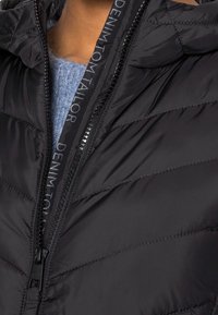 Černá prošívaná péřová bunda se zipovým zapínáním, prošívaným vzorem a páskem s logem podél zipu. Pod ním je viditelná měkká modrá textura.