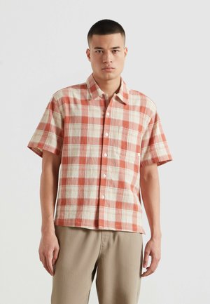 Mann trägt kurzärmeliges Hemd mit rot-weißem Kariertem Muster und beige Hose, steht vor einem einfarbigen hellen Hintergrund.